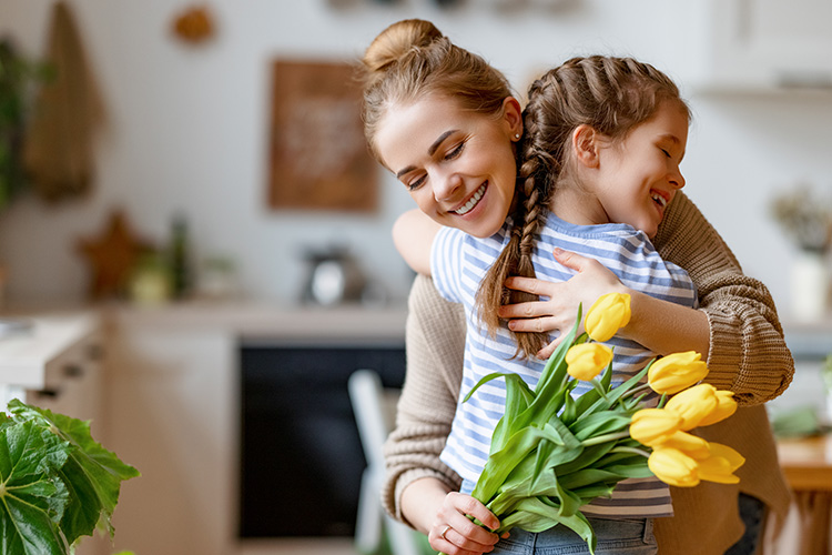 A mother and daughter hugging in the kitchen. Mom is holding a bouquet of yellow tulips.